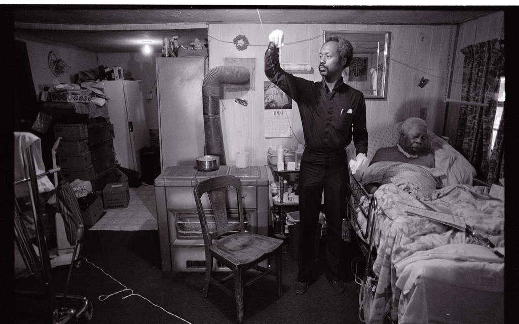 A man standing in the middle of a small room holds an object up to the light while an elderly man rests in a bed.