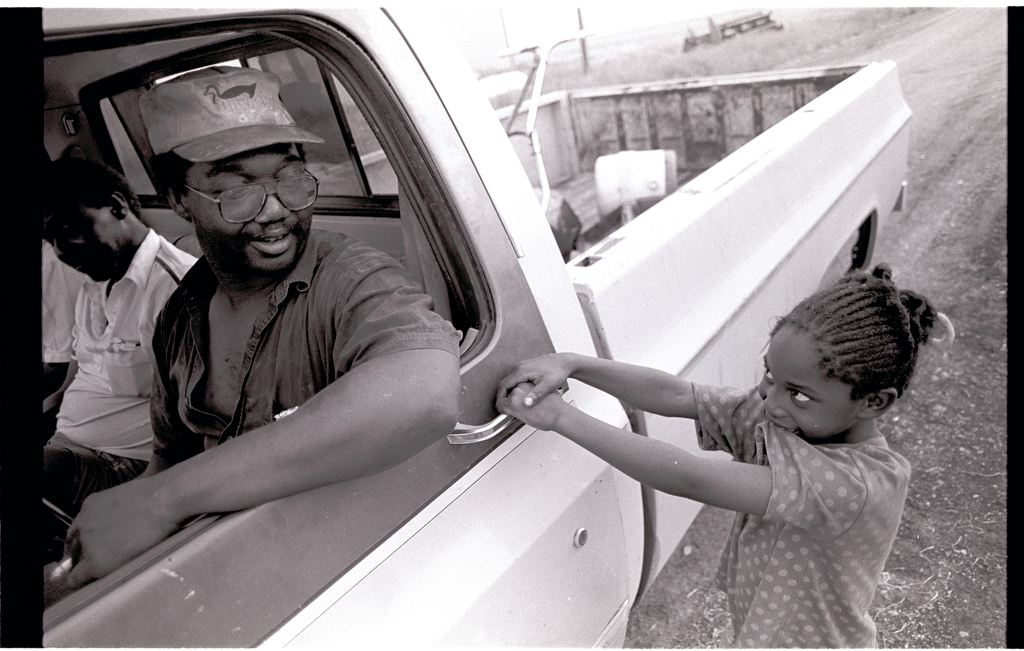 A young girl pulls on the door handle of a parked truck while the driver looks at her with a smile
