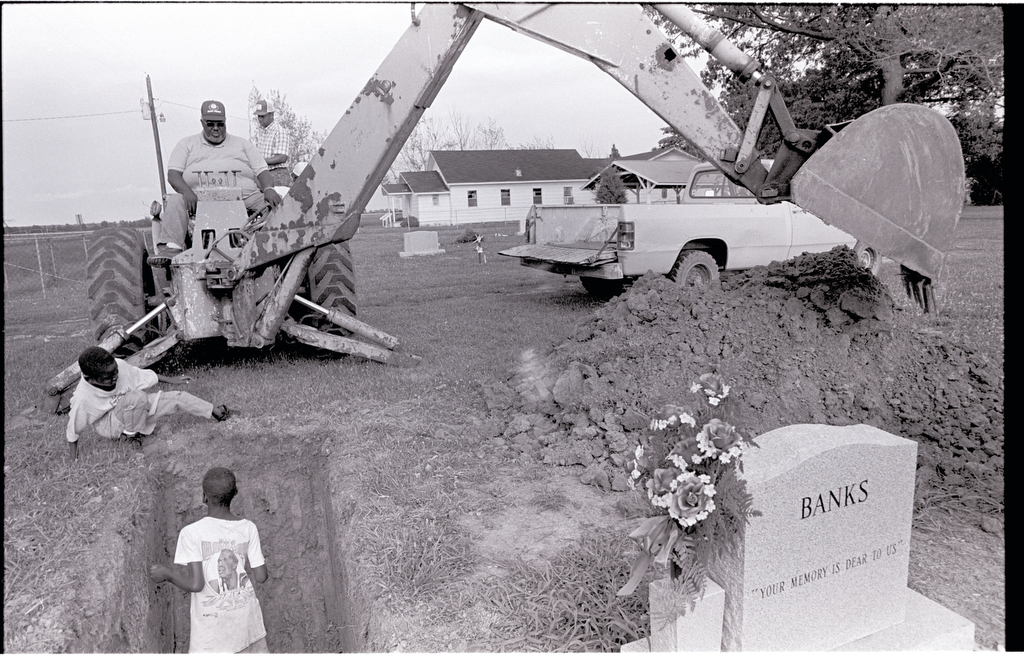 A man uses an excavator to dig a grave  as two young boys climb around the freshly dug grave. 