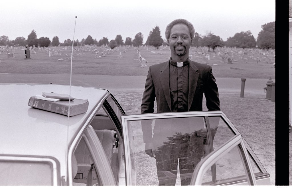 A man wearing the attire of a priest stands upright next to a car