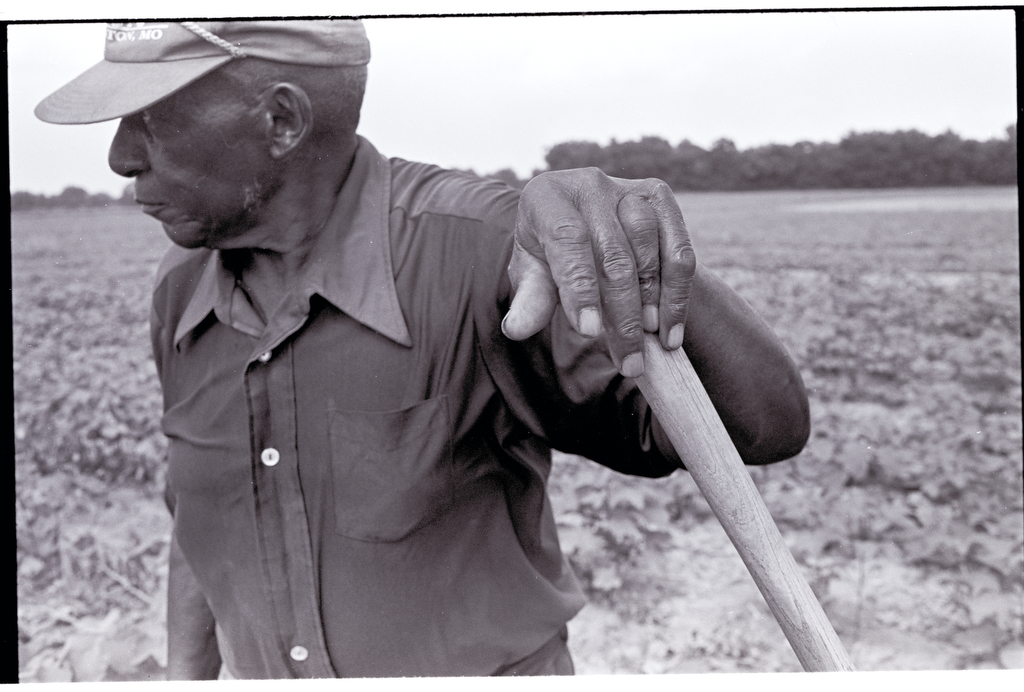 A farmer stands with his hand resting atop the handle of a tool while he looks out of frame.