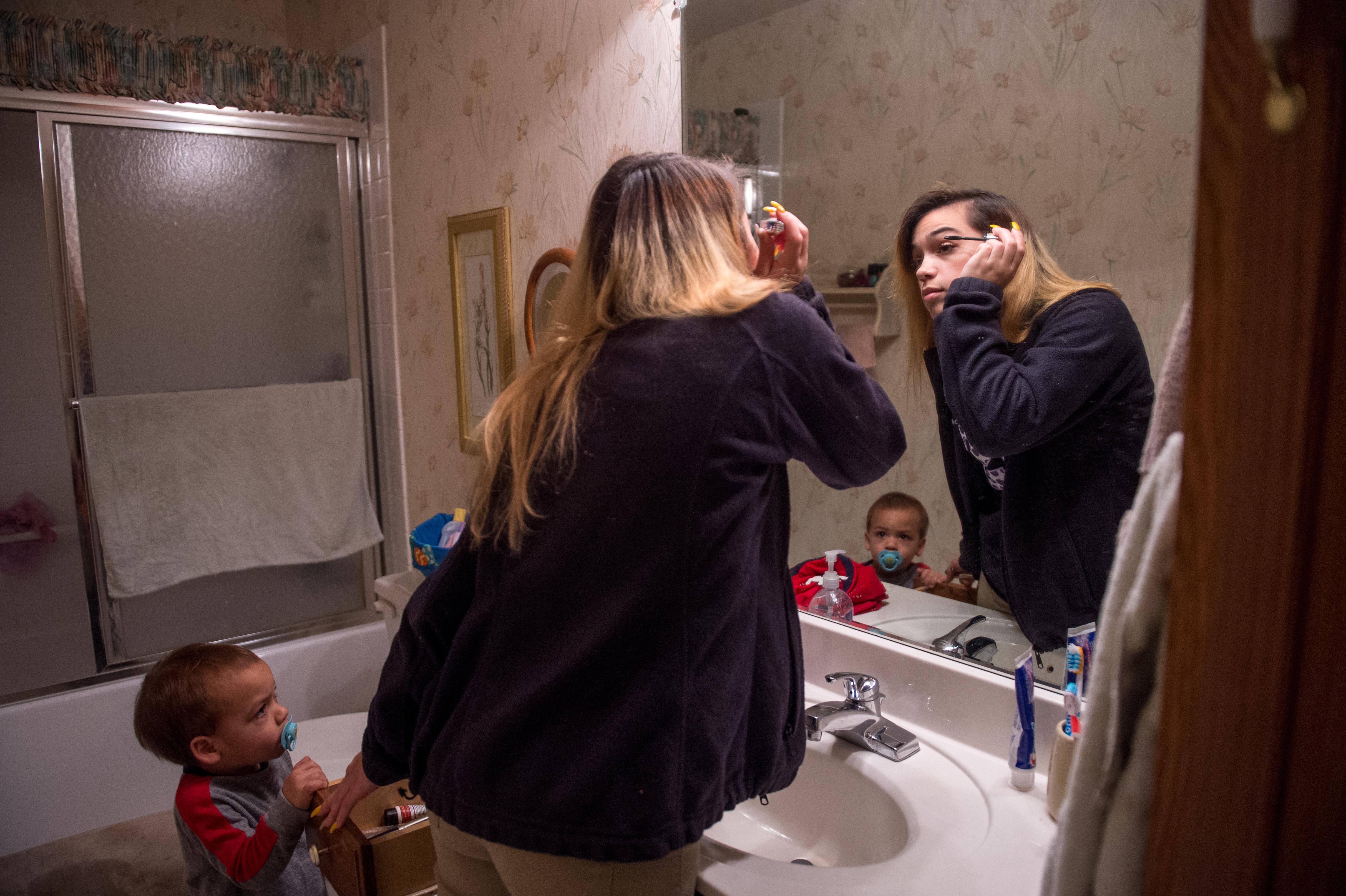 A young mother does her makeup in the bathroom as her child stands near by.