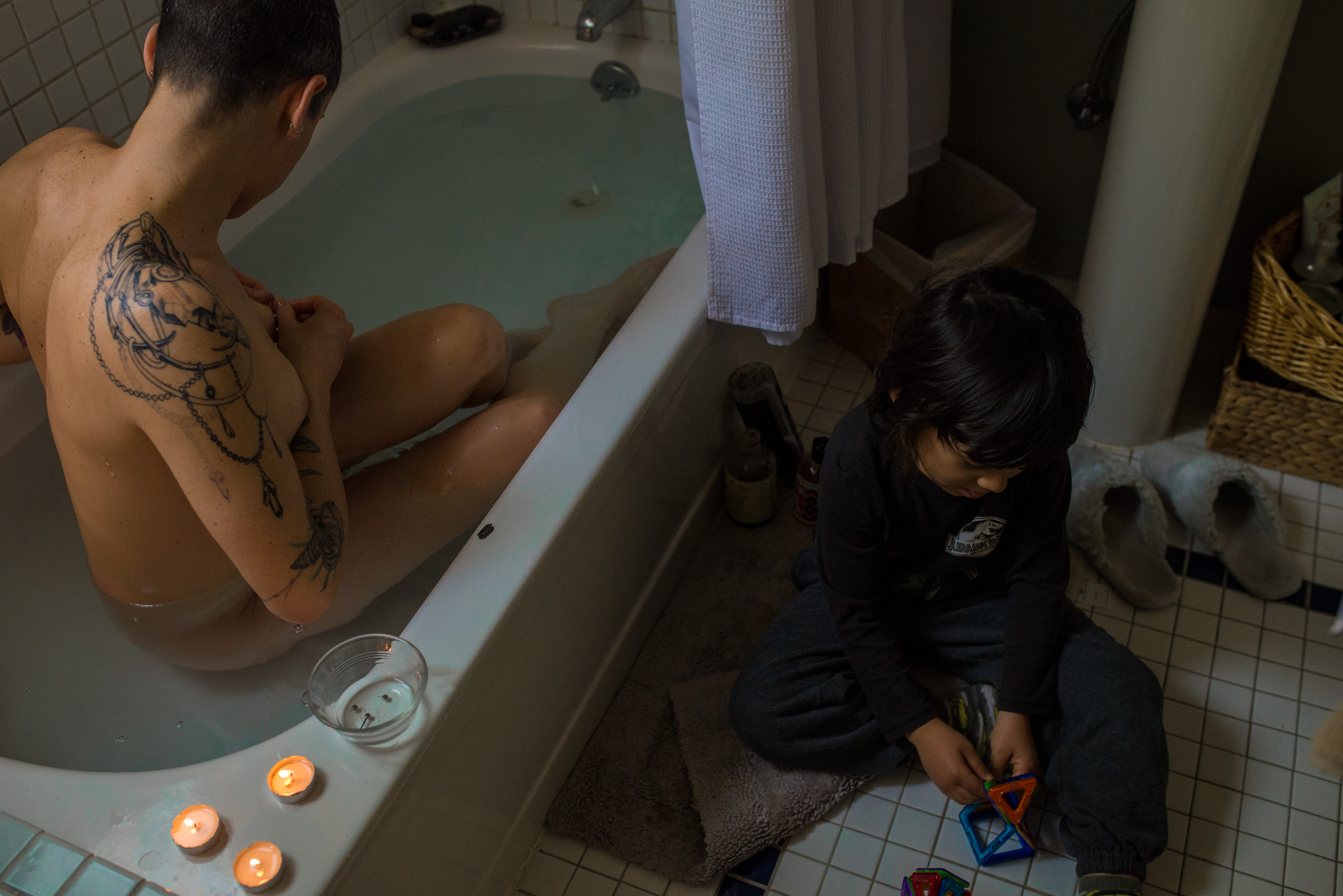 A woman takes a bath as a child plays with toys outside of the bath tub.
