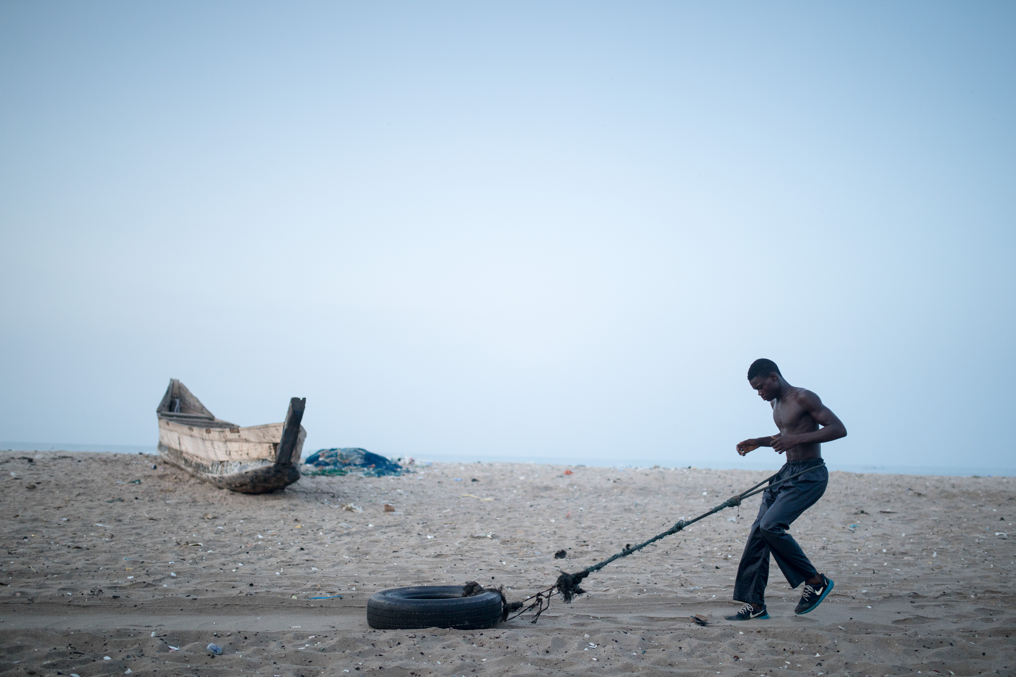 A young man runs backwards across the beach as he drags a tire tied to the end of a rope.