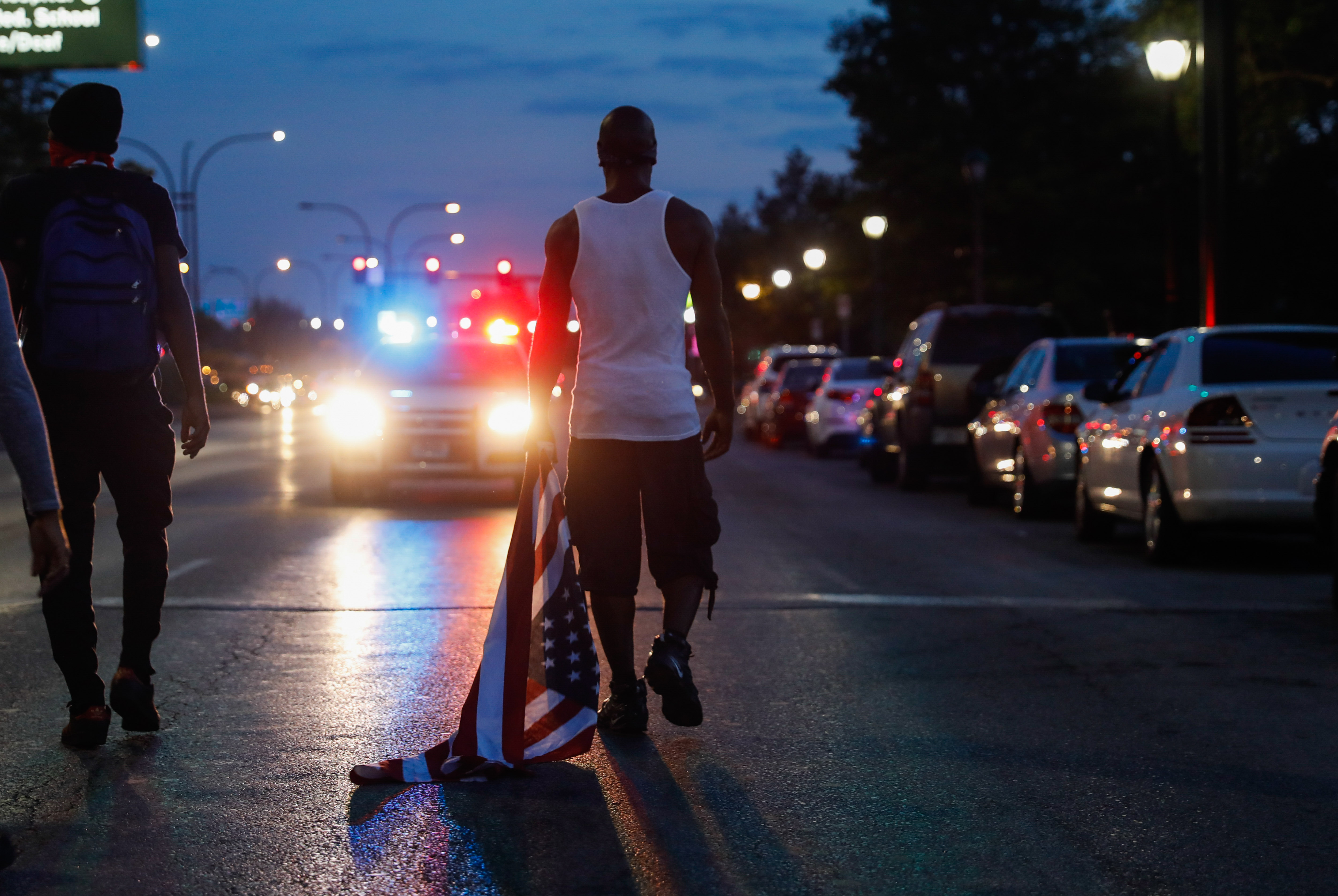A man taking part in a protest drags a U.S. flag behind him as he walks toward a parked police vehicle.