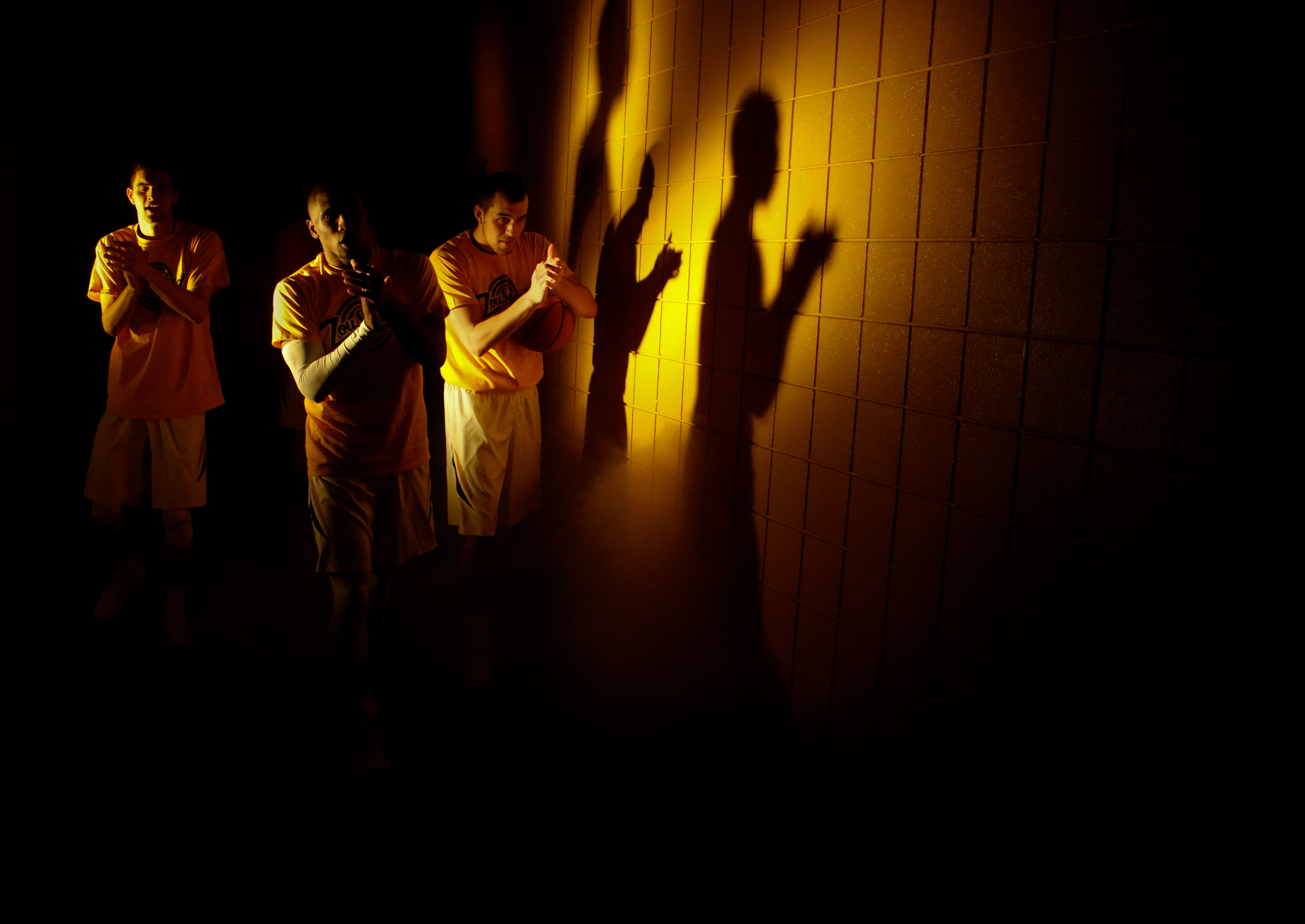 A trio of basketball players stand in beam of light casting their shadows against the wall.