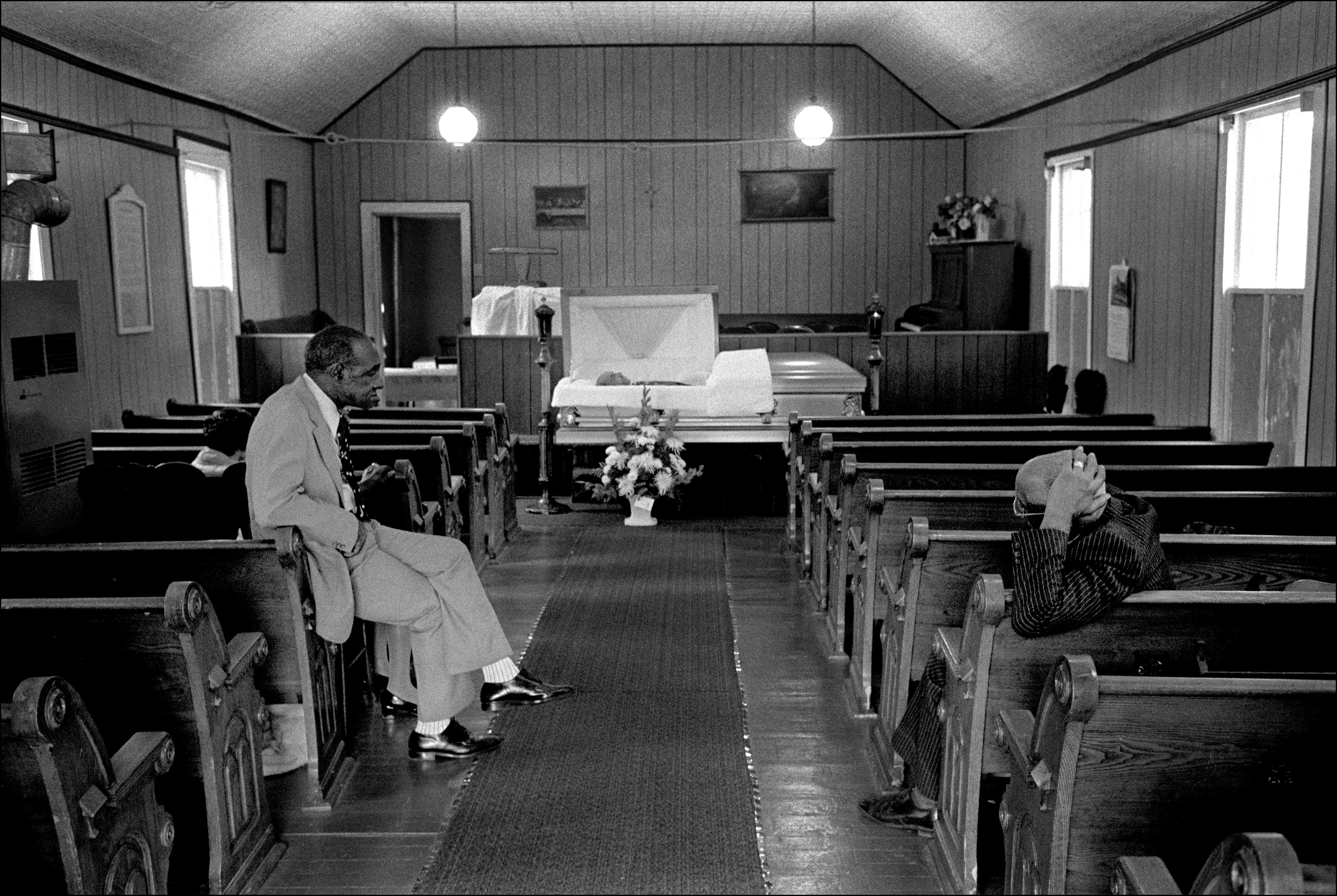 Two men sit across from each other in a chapel that is holding an open casket funeral.