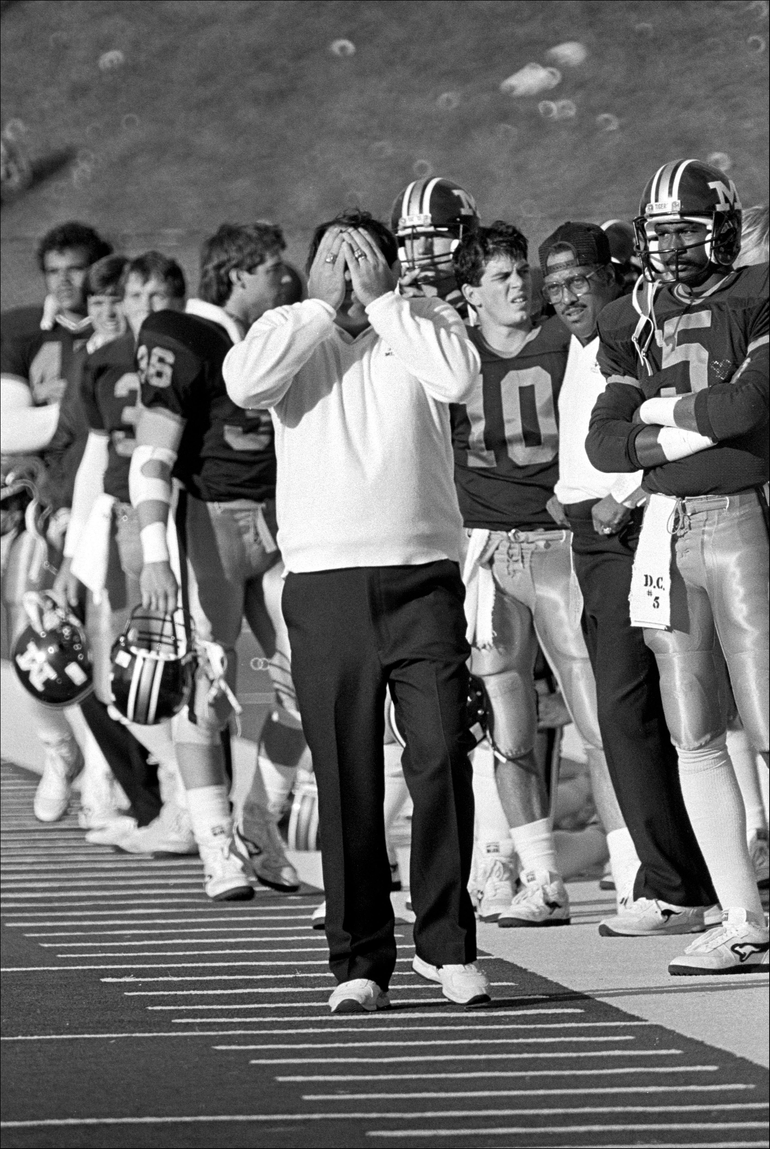 A football coach stands with his face in his hands along the sidelines during a game.