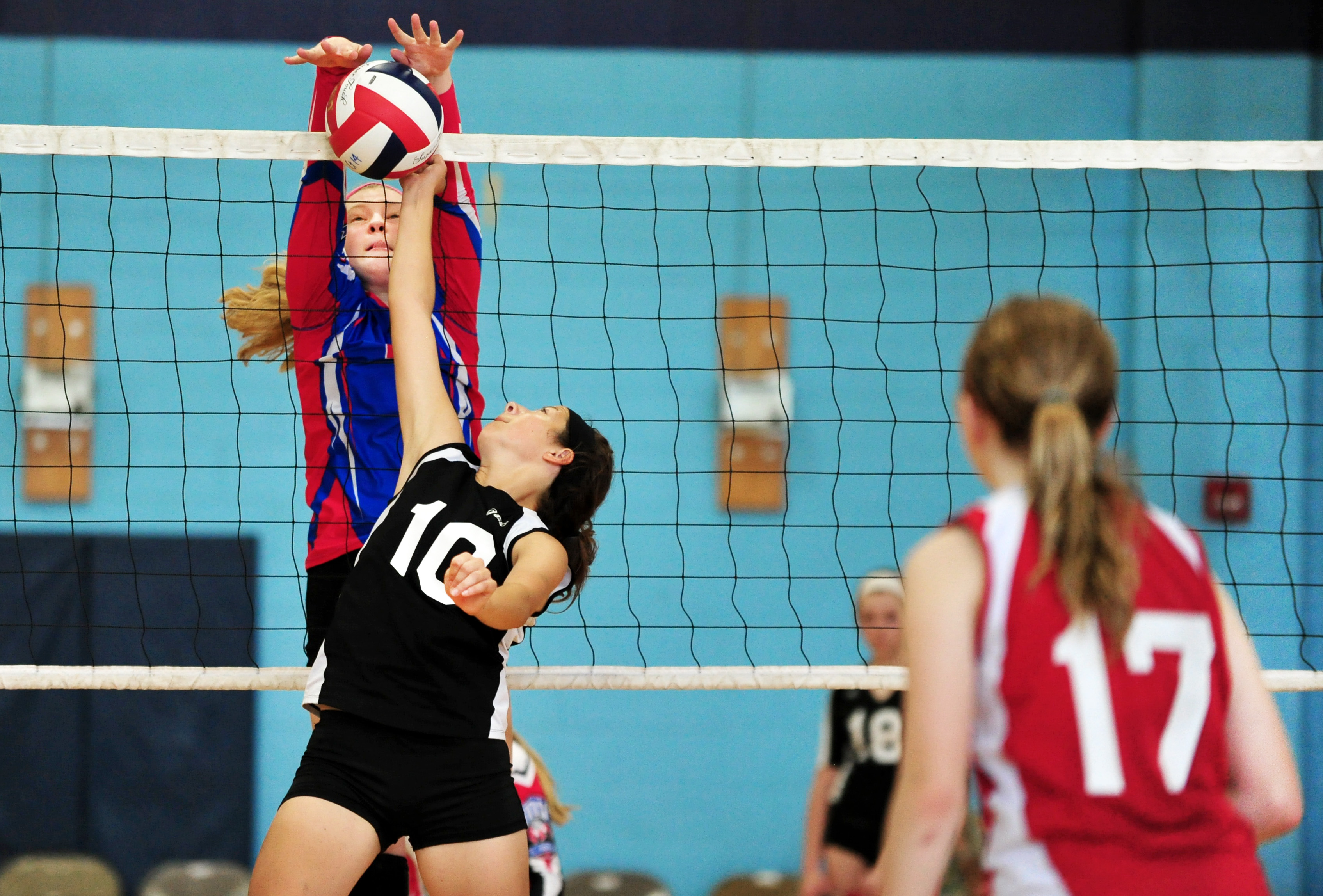 Volleyball players from opposing teams make contact with the ball at the net.