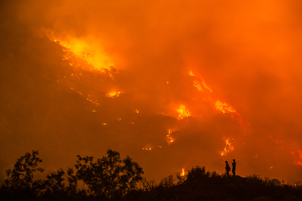 Two wilderness fire fighters stand silhouetted on a hill as a fire burns across the hills behind them.