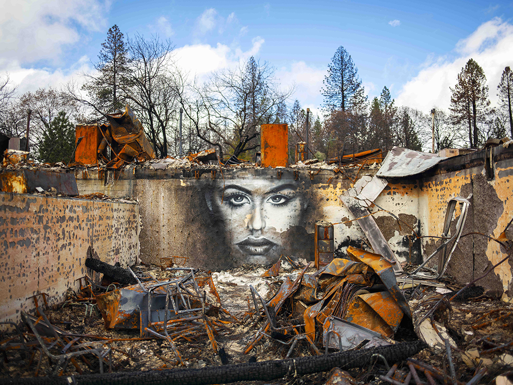 The remains of homes after being destroyed by a fire stand before a large mural of a human face.