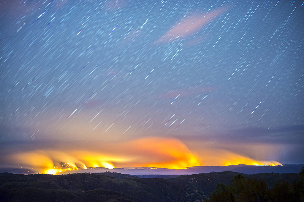 Trails of stars shine across the night sky as large fires burn along the horizon in the distance