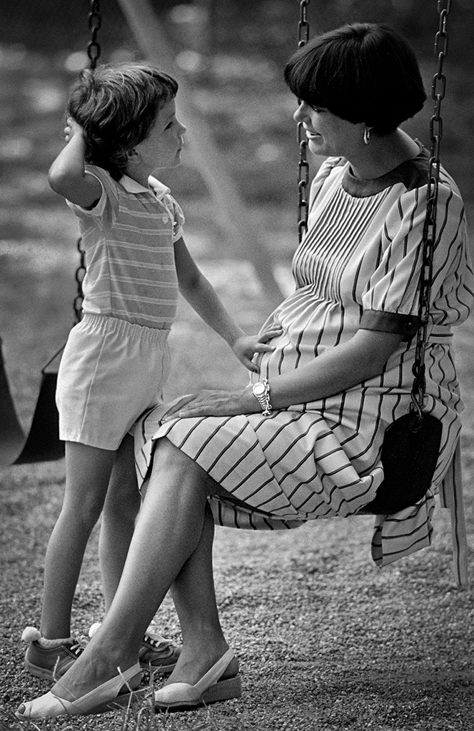 A pregnant woman sits on a swing at rest while talking to a child