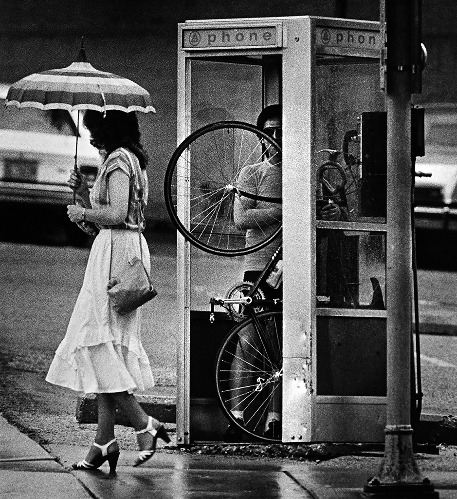 A person stands in a phone booth holds a bicycle standing vertically on the rear tire as a woman with an umbrella walks by.
