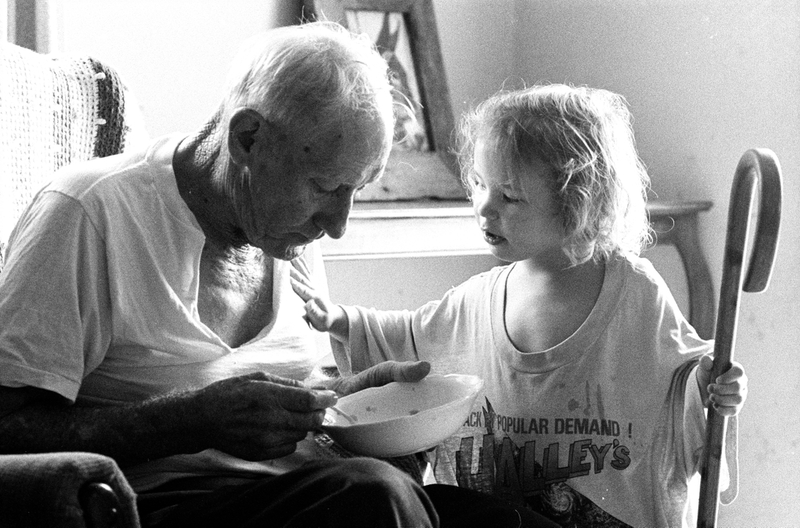 An elderly man eats from a bowl as a young child stands next to him.
