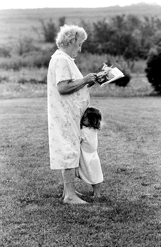 An elderly woman stands in field reading her mail as a young child presses their face against the elderly woman's legs.