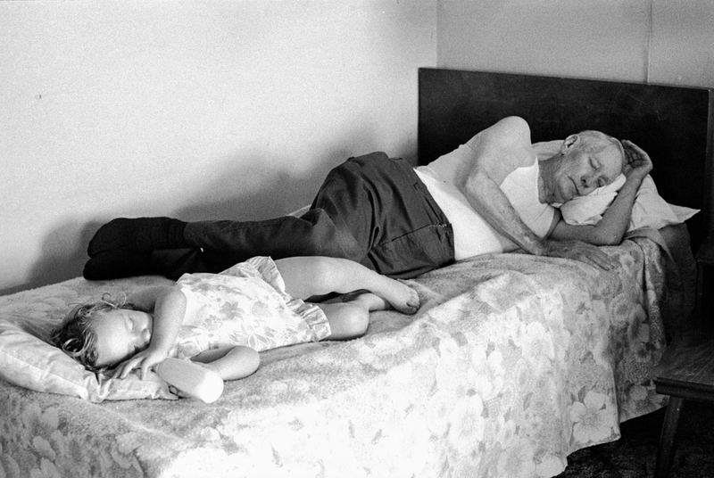 An elderly man and his granddaughter take a nap on a bed.