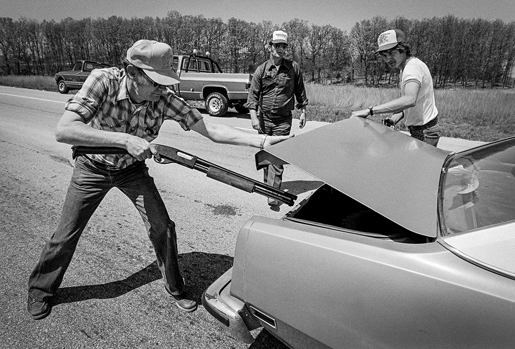 A trio of men, one holding a shotgun, inspect the trunk of a car.
