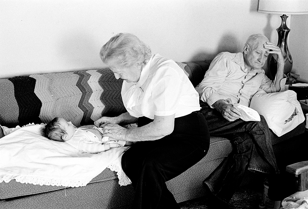 An elderly woman changes a baby's diaper on a couch as an elderly man sits at the end of the same couch.