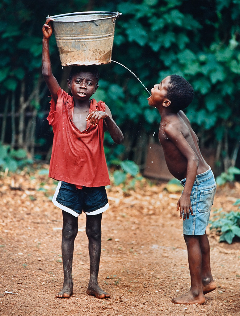 A boys holds a bucket of water atop his head while another boy drinks from a stream of water coming from a hole in the side of the bucket