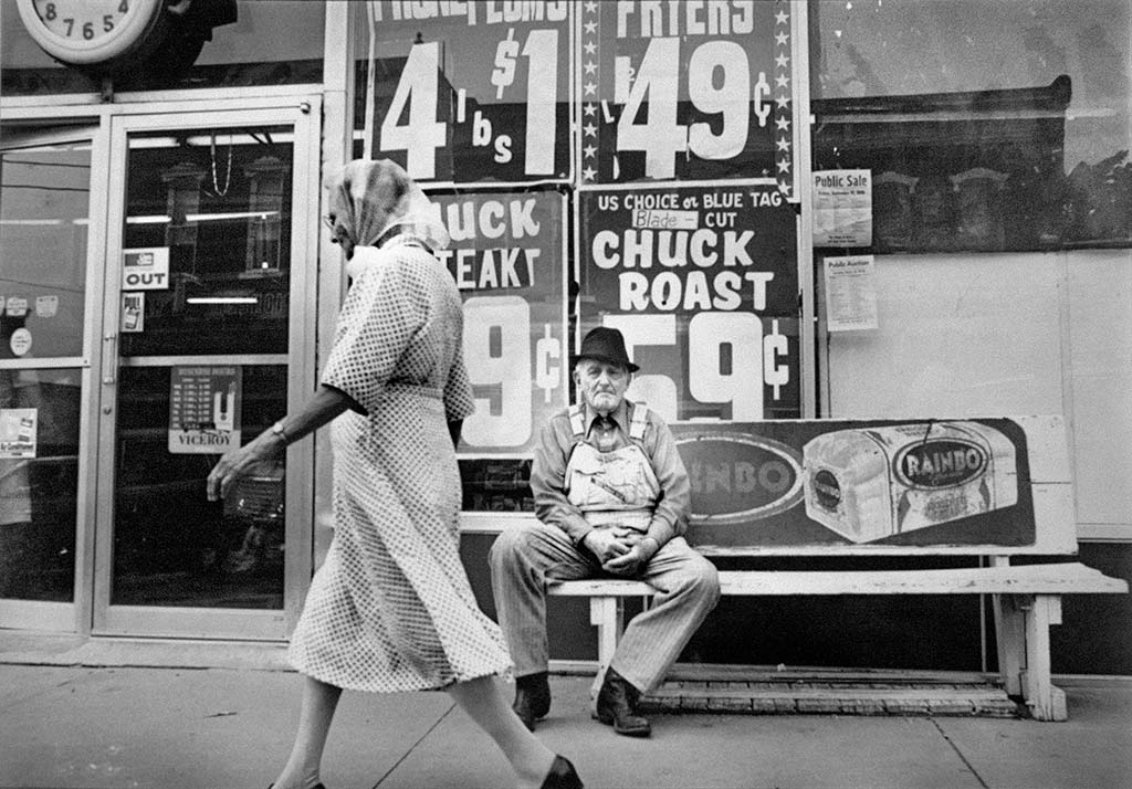 An elderly man sits outside of a grocery store as an elderly woman walks by.