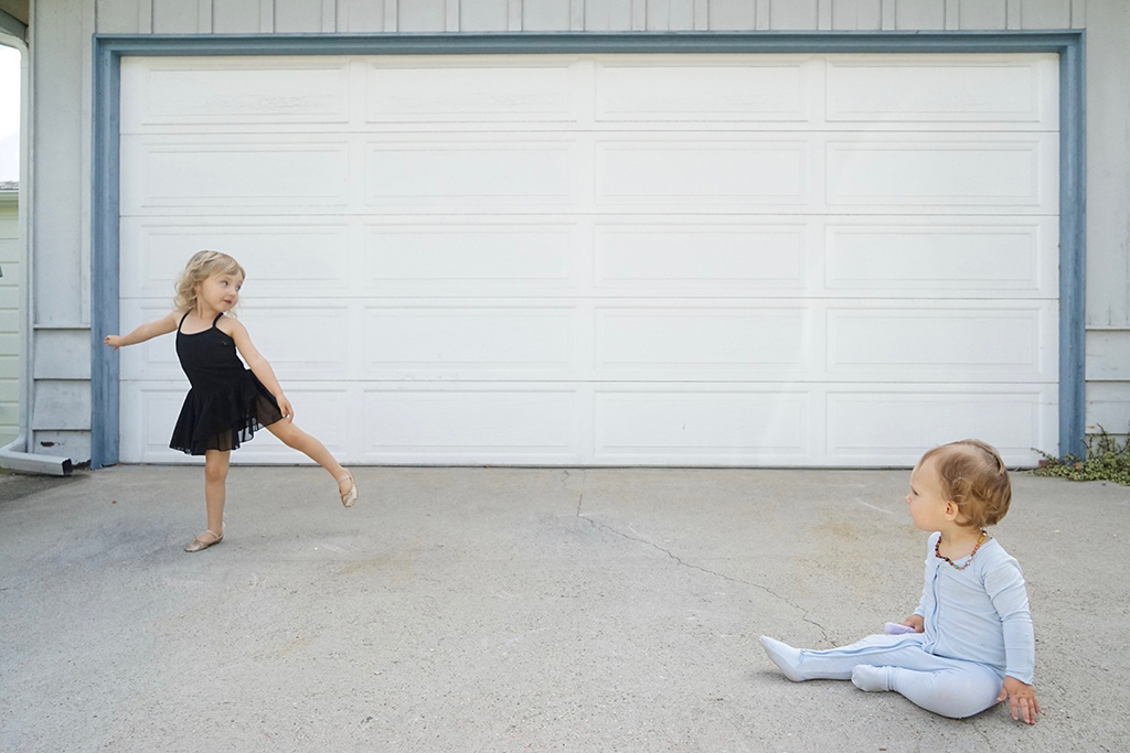 A baby sits on a driveway while a toddler dances about six feet away.