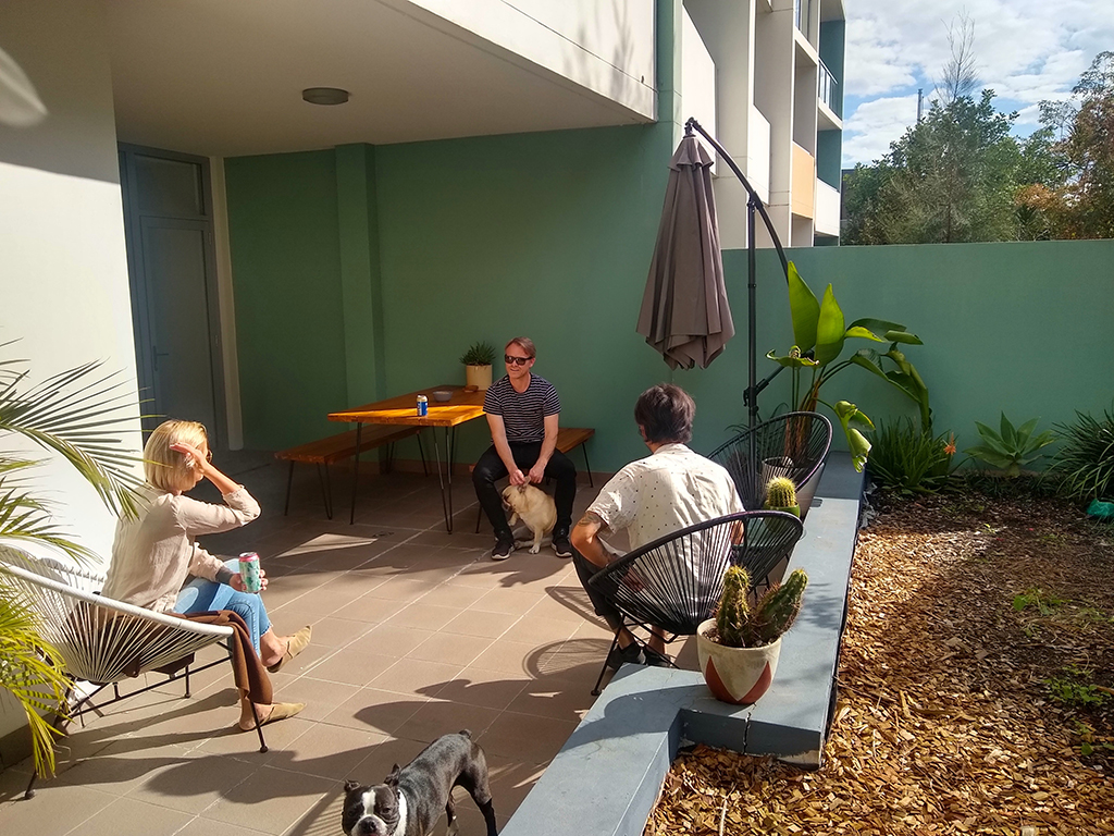 A trio of people practice social distancing while sitting on a patio. Two dogs can also be seen in the photo.