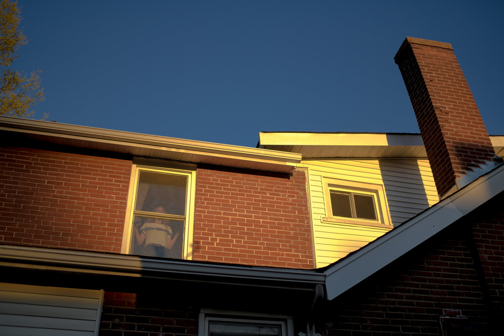 A person stands at a window of a house as seen from outside the house.