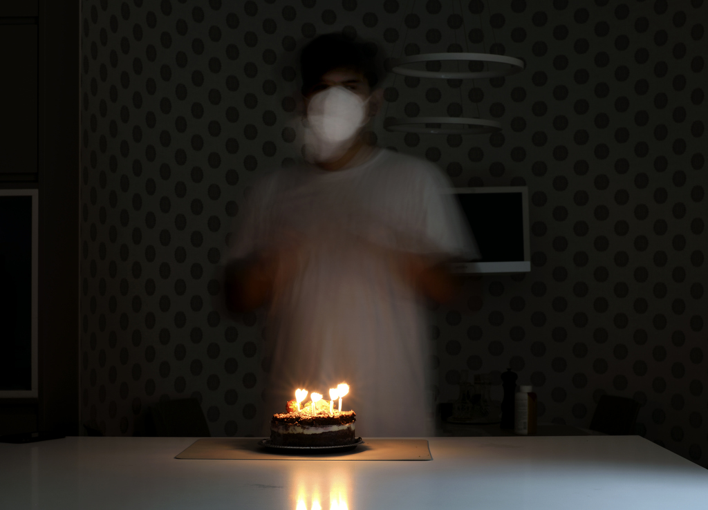 A person wearing a face mask stands before a cake with lit candles on it.