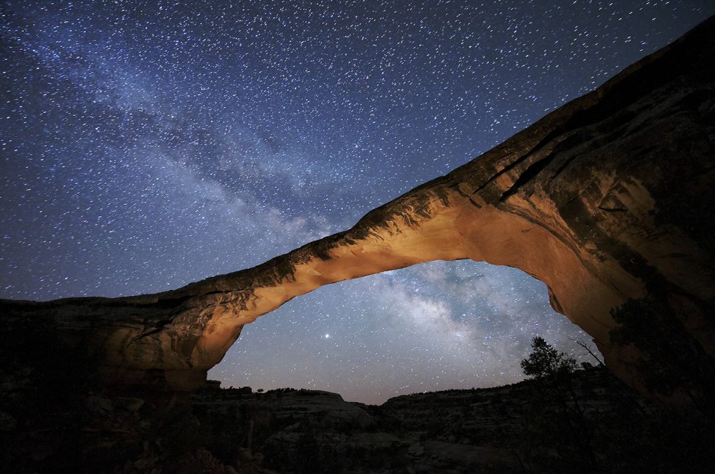 Night sky filled with bright stars shines over a natural bridge of rock.
