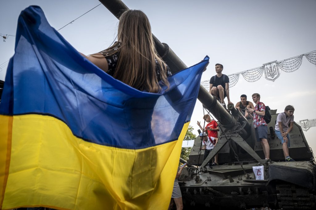 Woman holds up a Ukrainian flag while a group of young people climb on a parked military tank.