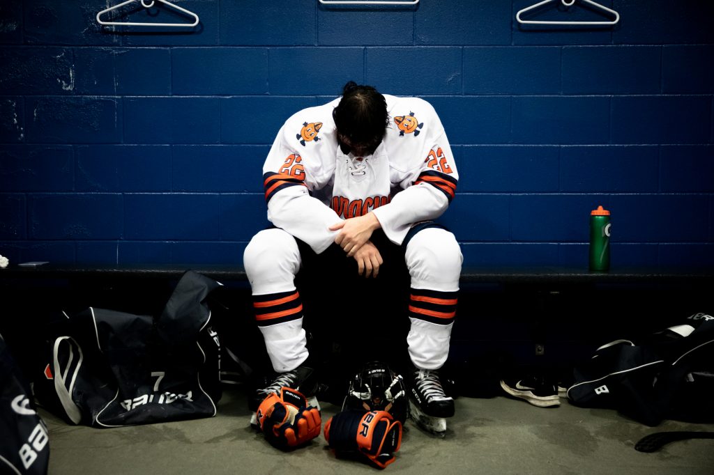 A hockey player hangs their head while sitting on a bench in a locker room.
