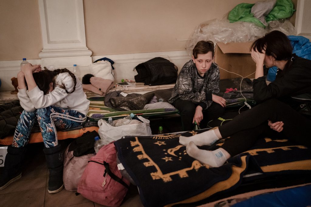Three displaced people sit on cots in a room among their belongs.