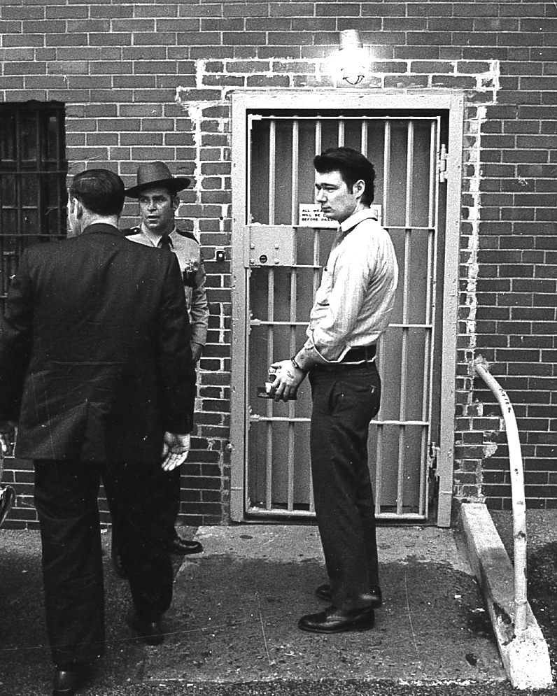 Man in handcuffs stands outside prison door with law enforcement officials.
