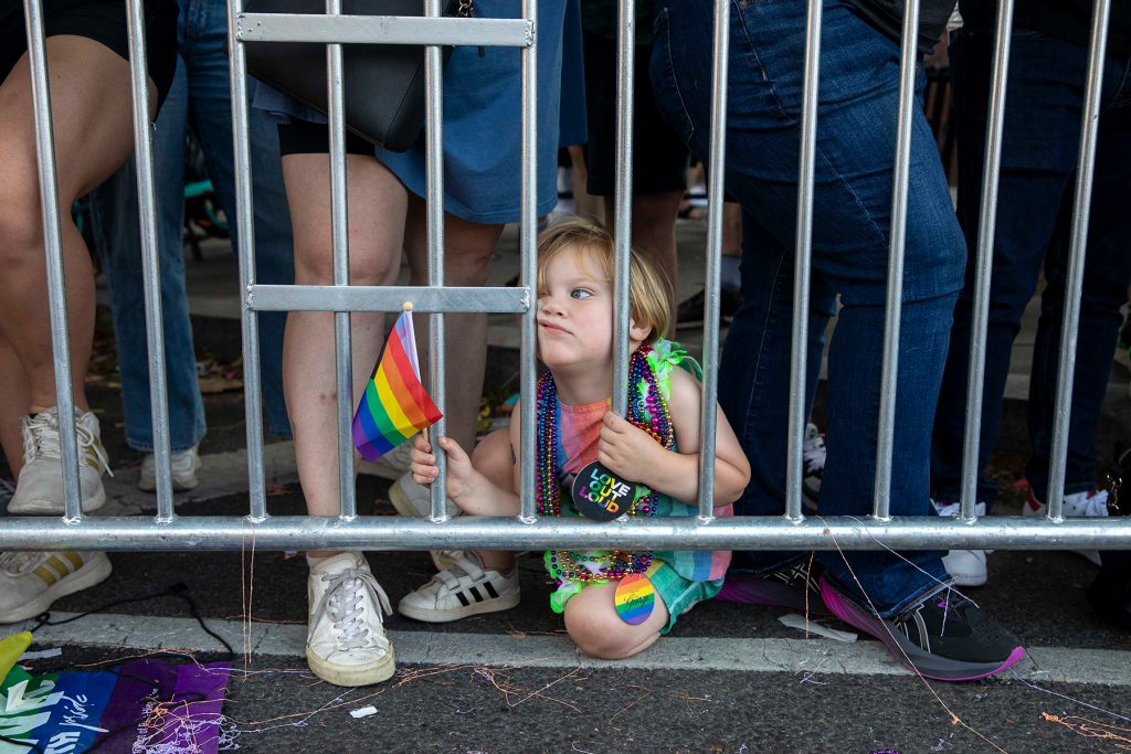 Girl sitting on the ground looking through a barricade and holding a pride flag
