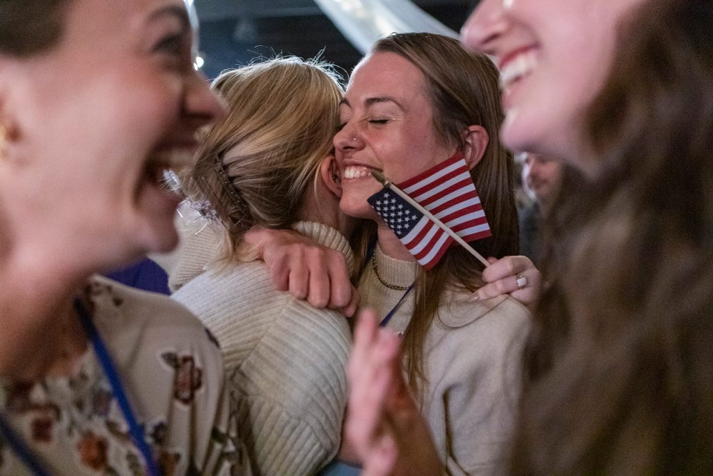 Photograph of two women hugging with other women surrounding them. One woman holds an American flag