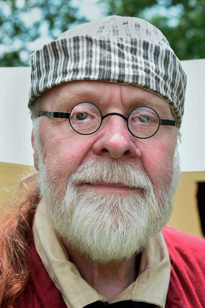 Photograph of an older bearded man wearing glasses, a red coat and a striped cap.