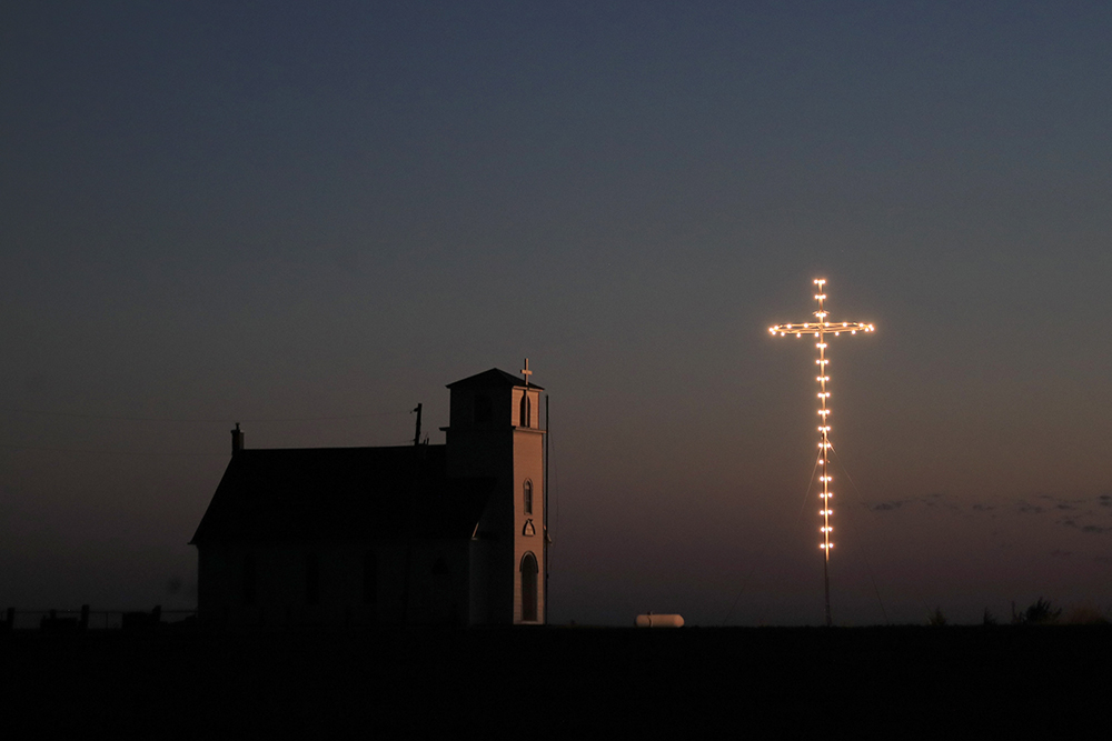 Photograph of a rural church with a lighted cross at dusk.