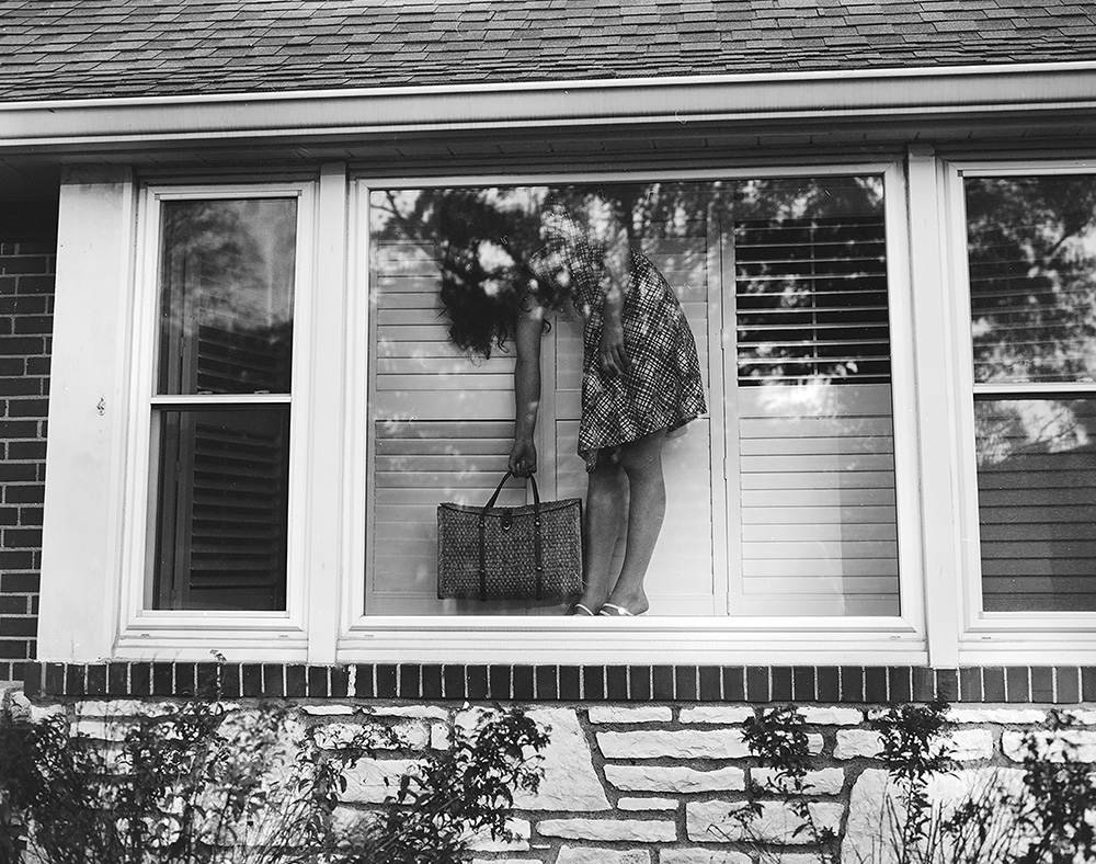 Photograph of a woman in a dress standing bent over in the window of a house. 